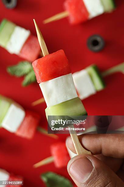 full frame image of unrecognisable person selecting mini bamboo kebab skewer with greek watermelon salad ingredients, alternating cubes of watermelon, feta cheese and cucumber, black olive slices, mint leaves, red background, elevated view - skewer stock pictures, royalty-free photos & images