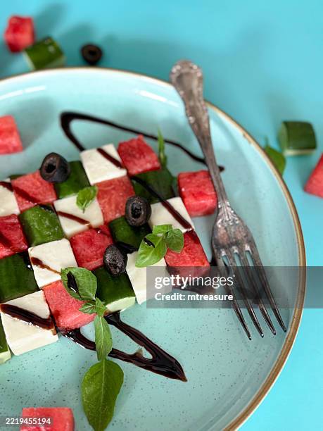 piatto blu di anguria insalata greca con cubetti alternati di anguria, formaggio feta e cetriolo, guarnita con fette di olive nere e foglie di menta, condita con aceto balsamico, forchetta di metallo, vista dall'alto, alimentazione sana - oliva nera foto e immagini stock