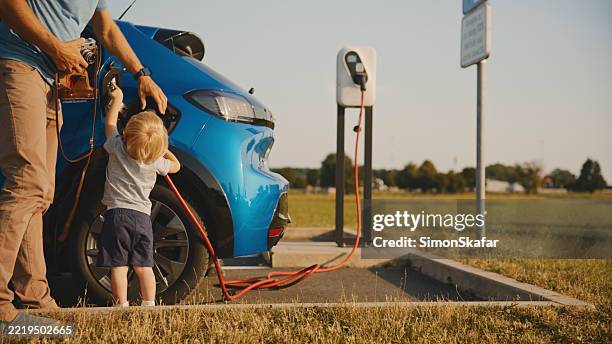 cute toddler son helping father with retro camera charge electric car at electric vehicle charging station - electric car stock pictures, royalty-free photos & images