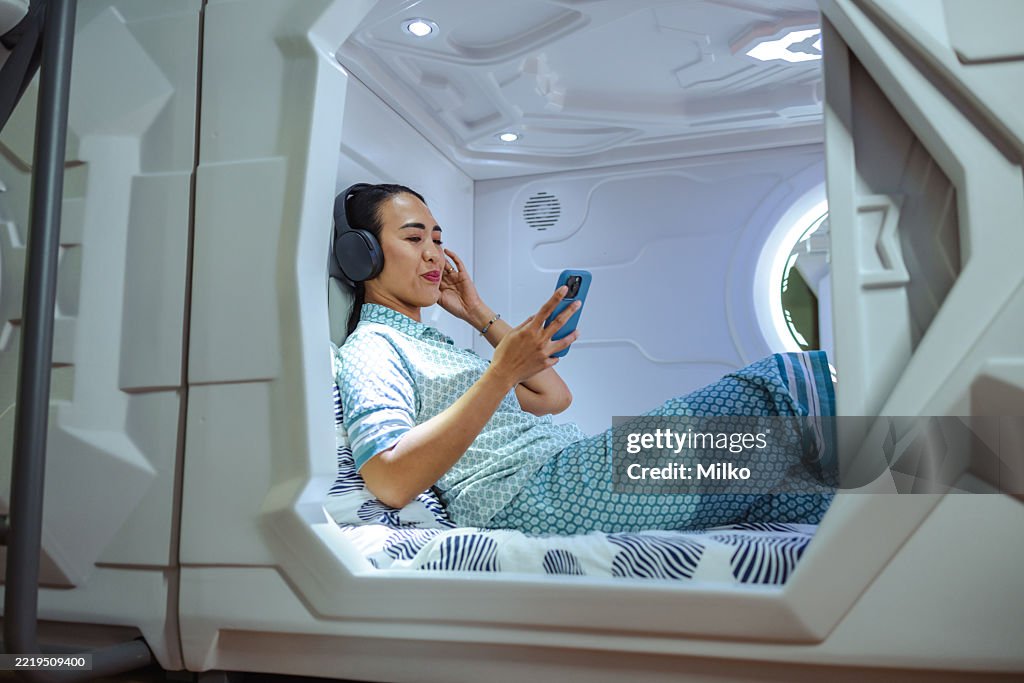Woman relaxing in modern sleep pod and consuming media on smartphone