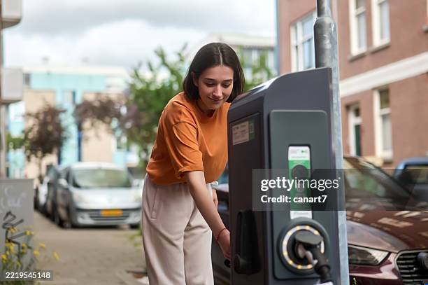 young woman plugging charging cable into electric car at public charging station - charging station stock pictures, royalty-free photos & images