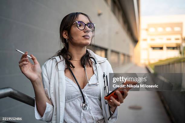 ärztin in der pause, die ein telefon hält und im freien in der nähe eines modernen gebäudes raucht - zigarettenpause stock-fotos und bilder