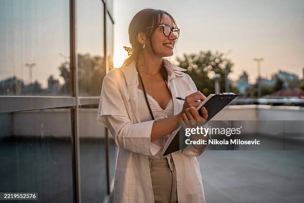 smiling female professional in sunlit urban setting with clipboard - golden hour stock pictures, royalty-free photos & images