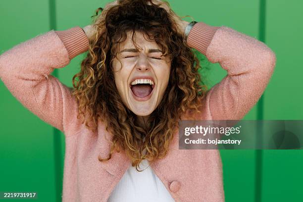 frustrated and angry woman is screaming out loud and pulling her hair on green background - histeria imagens e fotografias de stock