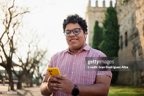 young man using smartphone and wearing earbuds in urban park - body positive stock pictures, royalty-free photos & images