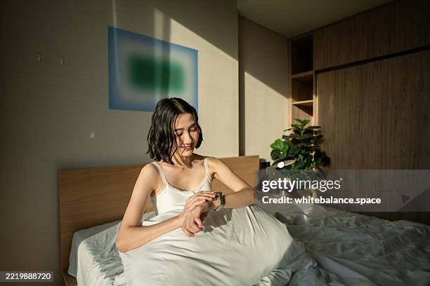 woman sitting and stretching on bed, looking out to the city skyline, tapping her smart watch to check notifications - fitness tracker stock pictures, royalty-free photos & images