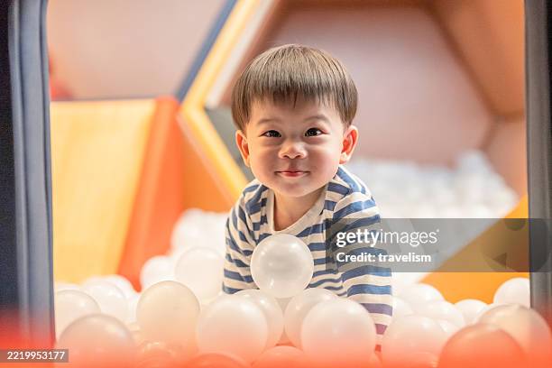 a playful two-year-old boy enjoys sliding into a vibrant ball pool at an indoor playground. with bright smiles and cheerful energy, the toddler explores the colorful environment, creating joyful moments of active play, sensory learning, - baby ball pool stock pictures, royalty-free photos & images