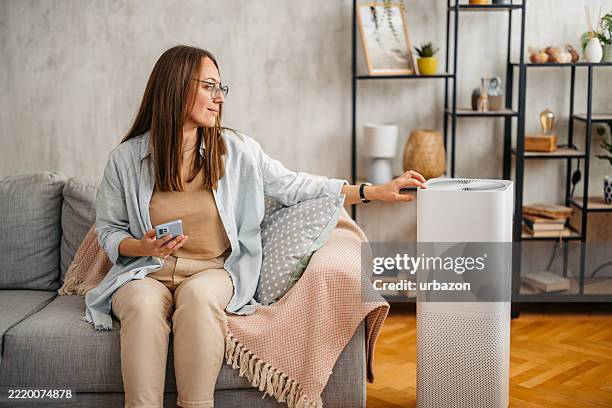 young woman turing on the air purifier using her phone while sitting on the sofa at home - air purifier stock pictures, royalty-free photos & images