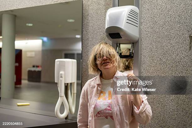 a blonde woman stands in front of a hand dryer in a modern public restroom - hand dryer stock pictures, royalty-free photos & images