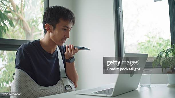 asian man with arm cast using voice assistant while working. - translation services stock pictures, royalty-free photos & images