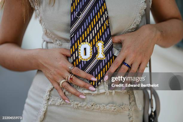 Gabriella Berdugo wears a sleeveless dress in a light beige color with frayed edges along the seams. The dress is paired with a striped tie featuring...