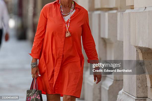 Passerby wears an orange long-sleeved shirt dress with a classic collar and button-down front. Layered colorful beaded necklaces are visible, one...