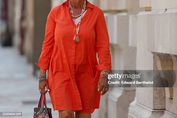 Passerby wears a bright orange shirt dress with a classic collar and button-down front. Layered multicolored beaded necklaces are visible, featuring...