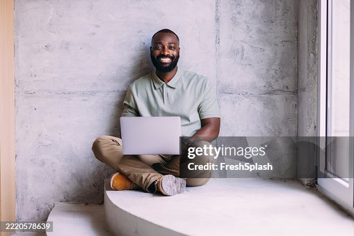 Smiling Man Sitting Indoors While Using Laptop in Modern Concrete Interior Smiling Man Sitting Indoors While Using Laptop in Modern Concrete Interior