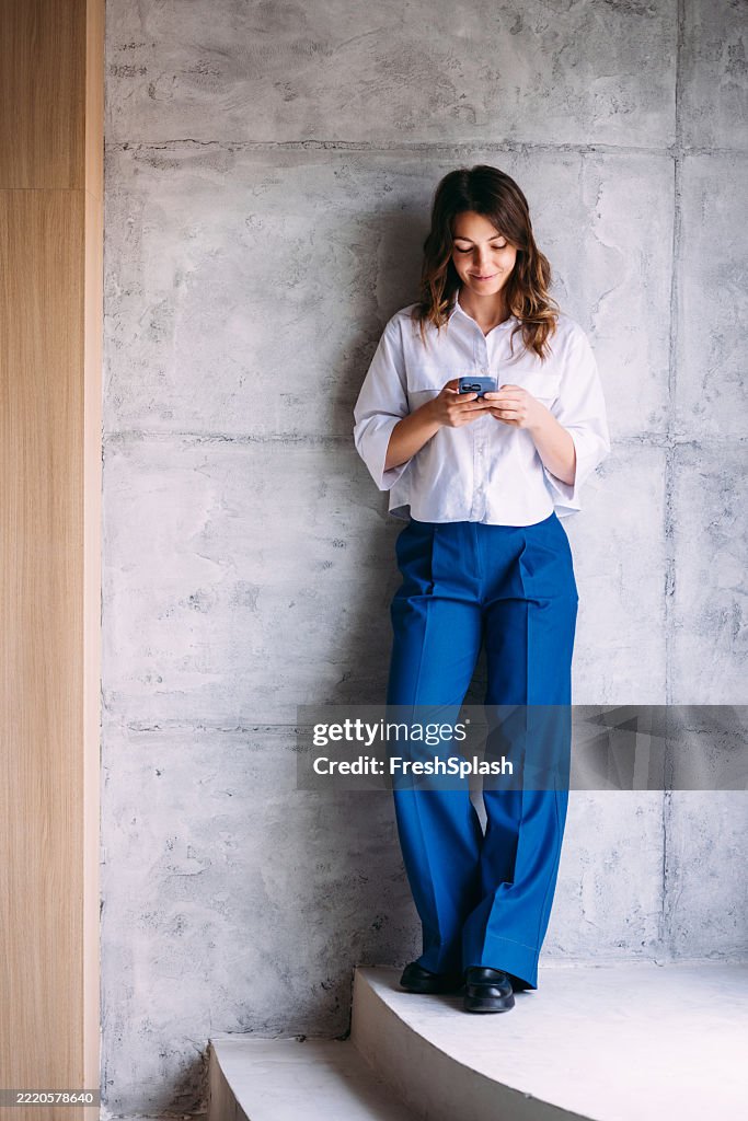 Stilvolle Frau In Blauen Hosen Checkt Ihr Telefon, Während Sie Drinnen Lächelt