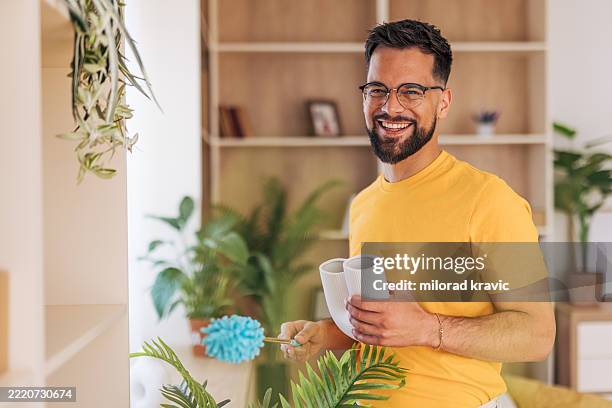 young man dusting houseplants and holding ceramic vases, smiling at camera - man dusting stock pictures, royalty-free photos & images