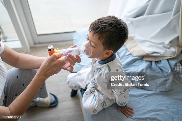 child using breath spacer with an aerosol inhaler with caregiver in soft bedroom light. - asthmatic stock pictures, royalty-free photos & images