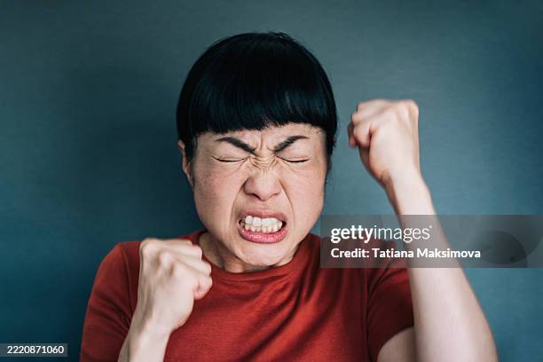 angry asian woman clenching fists and tightly closing eyes. concept of inner rage, frustration, and emotional outburst under pressure. - kwaad stockfoto's en -beelden
