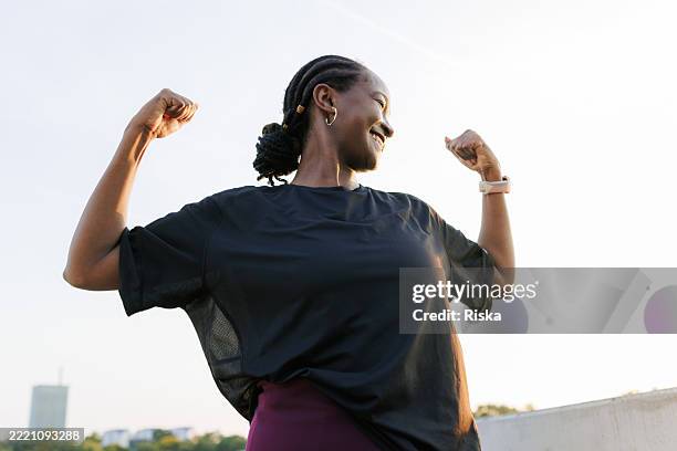 mujer negra segura de sí misma flexionando los músculos y sonriendo bajo un cielo despejado - flexionar los músculos fotografías e imágenes de stock