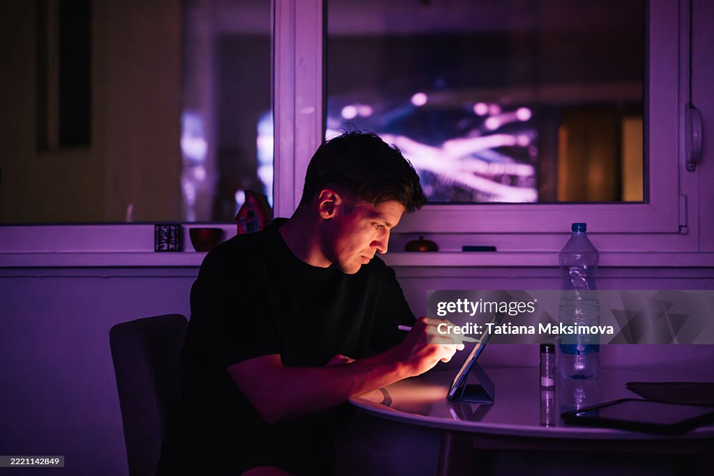 Man working at a tablet in a dark room illuminated by a purple glow from the screen and window reflections.