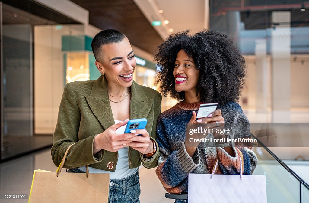Two smiling women shopping online using a smartphone and credit card in a mall
