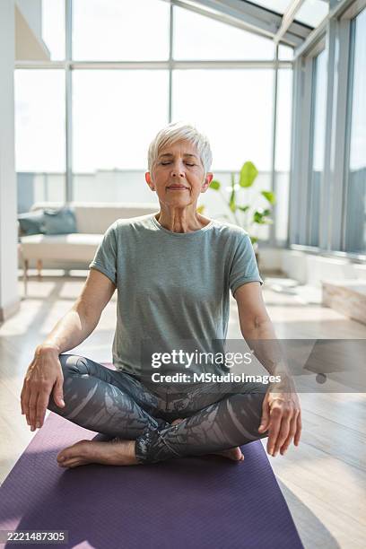 senior woman practicing yoga in a bright sunlit living space - lotus position stock pictures, royalty-free photos & images