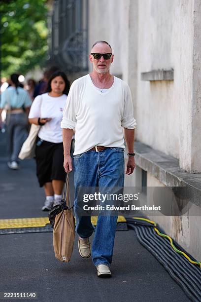 Guest wears black sunglasses and a white V-neck sweater with the sleeves rolled up. Underneath, a white undershirt is visible. Blue denim jeans are...
