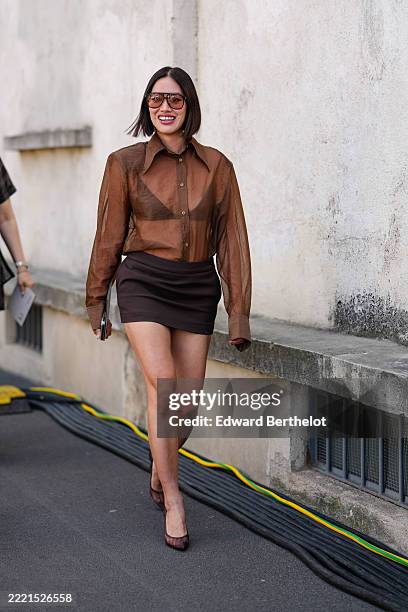 Tiffany Hsu wears dark brown sunglasses, light brown buttoned up sheer mesh long sleeve shirt, black bralette, dark brown mini skirt, dark brown...