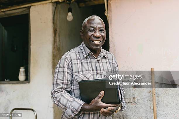 anciano sosteniendo la biblia y sonriendo cerca de la pared rústica - países en vías de desarrollo fotografías e imágenes de stock