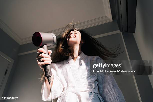 a young woman in white bathrobe blow-dries her hair at home. - hair dryer stock pictures, royalty-free photos & images