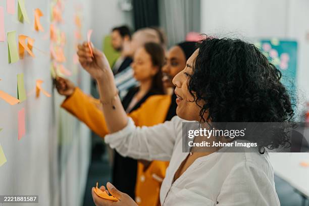 smiling curly haired businesswoman sticking adhesive note on whiteboard in office - scandinavie photos et images de collection
