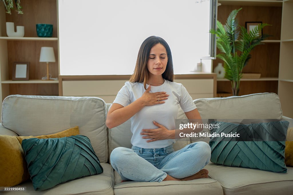 Serene woman practicing deep breathing exercise on sofa at home