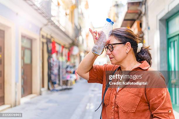 mature woman suffering from heat stroke in the city, drinking water and touching her forehead. heat wave in summer sunny days, eyes closed and hydrating - temperatura imagens e fotografias de stock