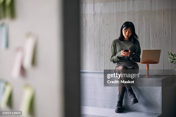 female entrepreneur using smart phone while sitting on bench next to laptop at office - de pernas cruzadas imagens e fotografias de stock