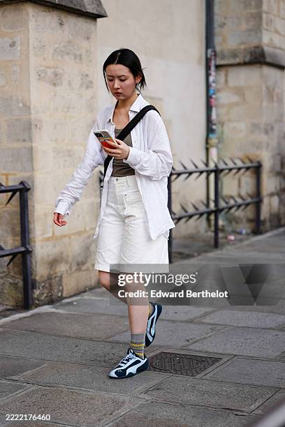 Guest wears a white, long-sleeved, button-up shirt over a taupe tank top. The shirt is layered over white knee-length shorts with visible pockets and...