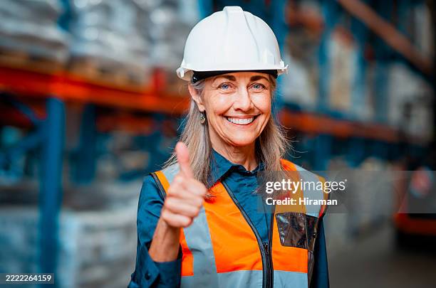 lavoratrice fiduciosa in un magazzino moderno che dà il pollice in su mentre indossa l'equipaggiamento di sicurezza e l'elmetto - attrezzatura di sicurezza foto e immagini stock