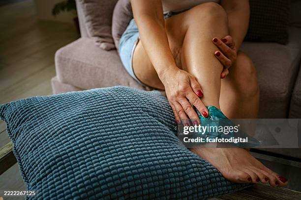 woman sitting on sofa applying cold gel pack on a painful ankle, arthritis or injury, ligament or cartilage problems. body care at home - swollen stock pictures, royalty-free photos & images