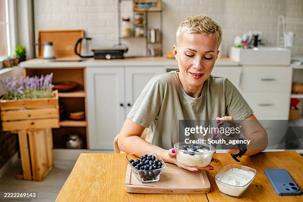 la mujer disfruta de un tazón saludable de arándanos mientras está sentada en una mesa de madera en una cocina moderna y luminosa - preparación de alimentos fotografías e imágenes de stock