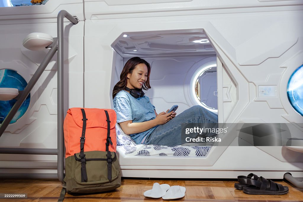 Young traveler relaxing in a modern capsule hostel room using a smartphone