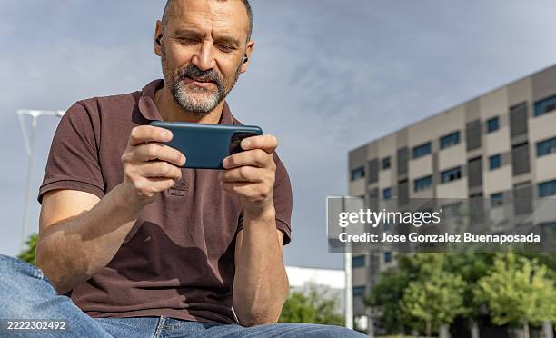 mature man playing mobile game using wireless earphones outdoors - spanish and portuguese people stock pictures, royalty-free photos & images