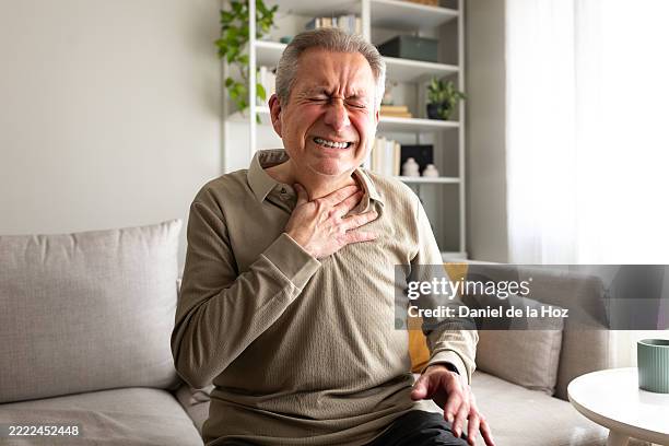 senior man suffering from chest pain while sitting on sofa at home - hartinfarct stockfoto's en -beelden
