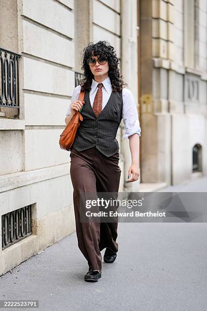 Loic Hornecker wears curly dark hair styled naturally, paired with large round sunglasses. A white button-up shirt with rolled-up sleeves is layered...