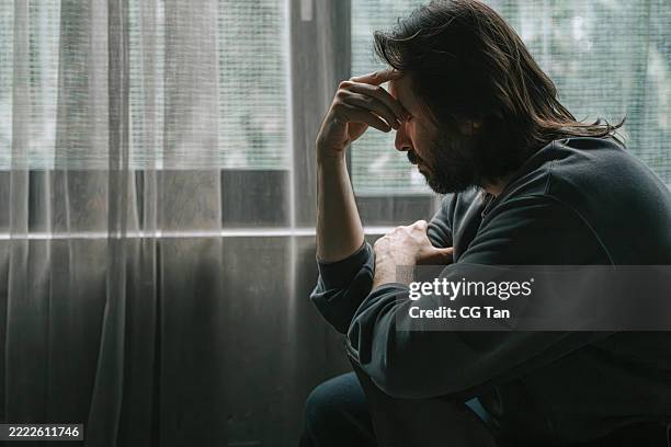 melancholic man sitting by window in deep thought - midlife crisis stock pictures, royalty-free photos & images