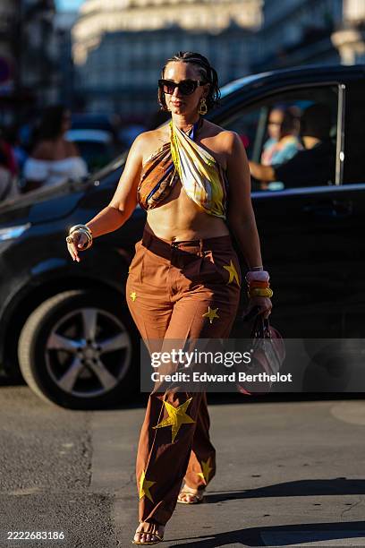 Guest wears black sunglasses, gold earrings, dark yellow dark brown pattern/print wrapped cropped scarf shirt, dark brown high waisted dark yellow...
