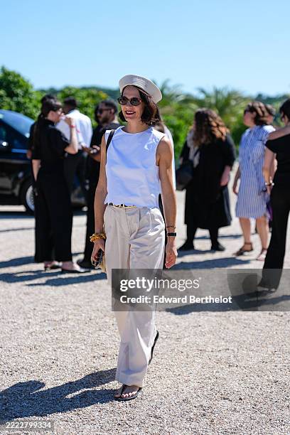 Guest wears a white sleeveless top with a scalloped hem, paired with high-waisted, wide-leg beige trousers. A beige beret is worn, and dark...