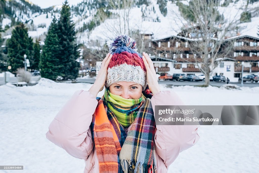 Caucasian woman wearing knit hat with pompom, colorful scarf, and pink jacket. Standing in snowy town with evergreen trees and buildings in background.
