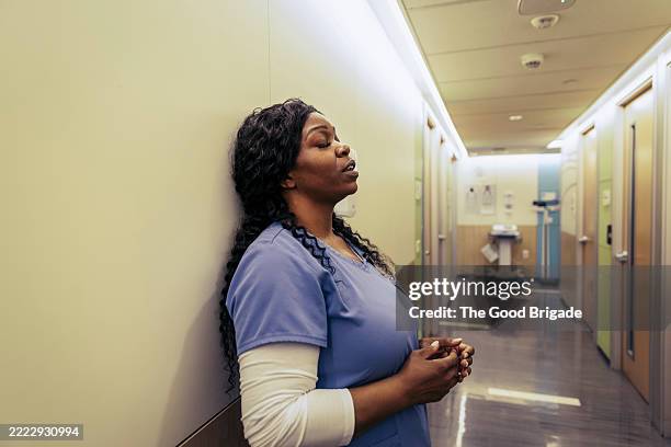 tired female nurse leaning on wall in hospital corridor with eyes closed - tired healthcare worker stock pictures, royalty-free photos & images