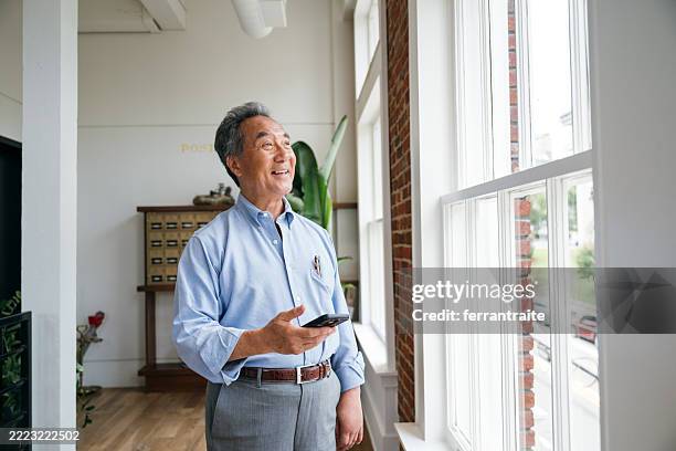 senior businessman portrait looking through the window while holding a smartphone - three quarter length stock pictures, royalty-free photos & images