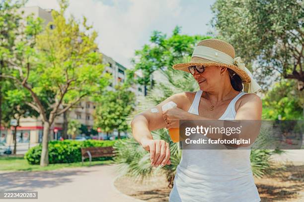 woman in casual clothing and wearing sunhat applying sunscreen on her arm. uv protection and skin care in the city. sunblock in a sunny summer day at the park - sun safety stock pictures, royalty-free photos & images
