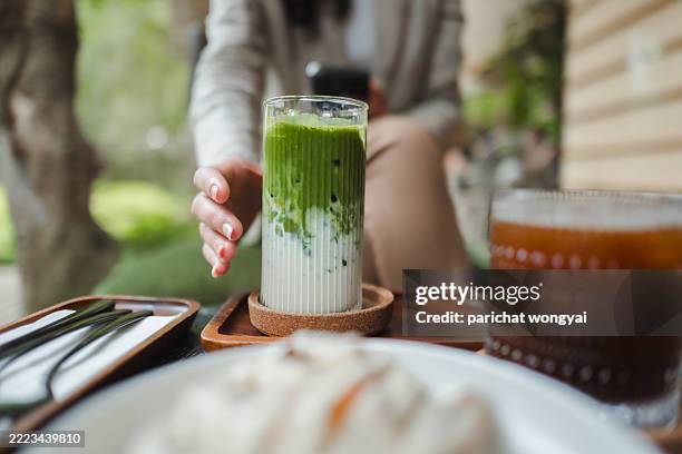 die hand greift nach einem erfrischenden eisgekühlten matcha-latte auf einem tisch, begleitet von einem glas eiskaffee und einem dessert, was eine gemütliche und einladende atmosphäre schafft - matcha stock-fotos und bilder